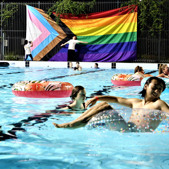 Group of people in a pool with floating rings, and a giant progressive pride flag in the background with a person standing in front of the flag arms stretched out being photographed by another person